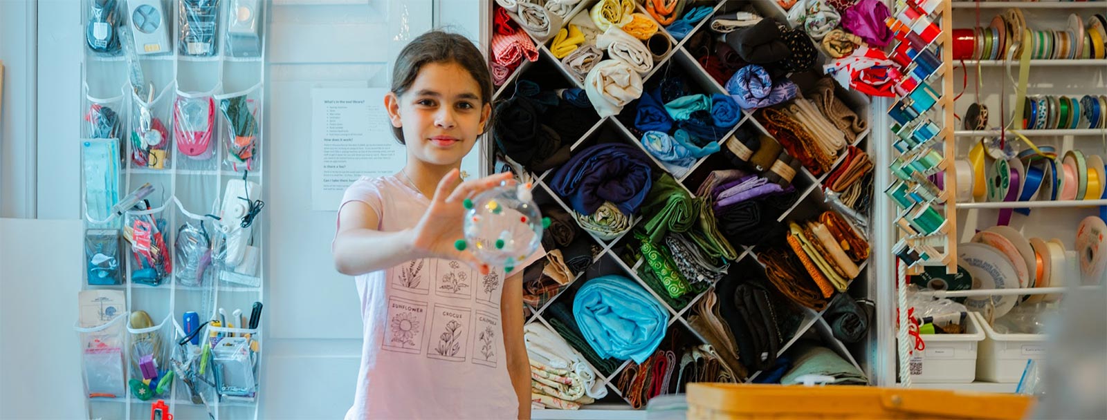 A girl holds up her art project in front of a wall of fabric and ribbons