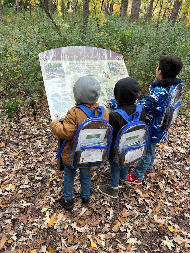 Three children consult a sign on the trail, wearing their Little Explorer's Backpacks