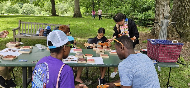 A child plays the ukulele, adults watching and assisting.