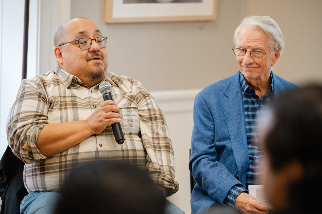 Two men sit at the front of the Great Room at Brushwood center. Jerry Loza holds a microphone.