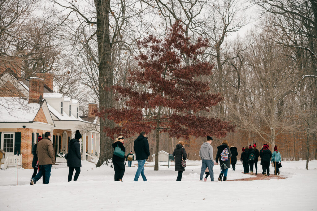 People in winter clothes walk along a path towards Brushwood Center. There is snow on the ground, and most of the trees are leafless. The sky is grey, but bright.